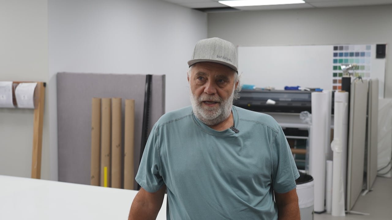 A man with a beard and a baseball cap with rolls of wallpaper in the background.