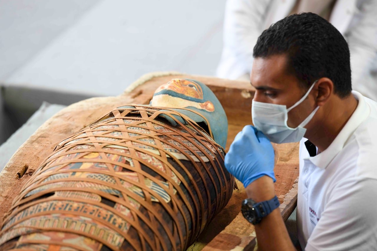An archaeologist inspects an Egyptian mummy that is wrapped in an elaborate and colourful burial shroud. The archaeologist is wearing a medical mask and gloves.
