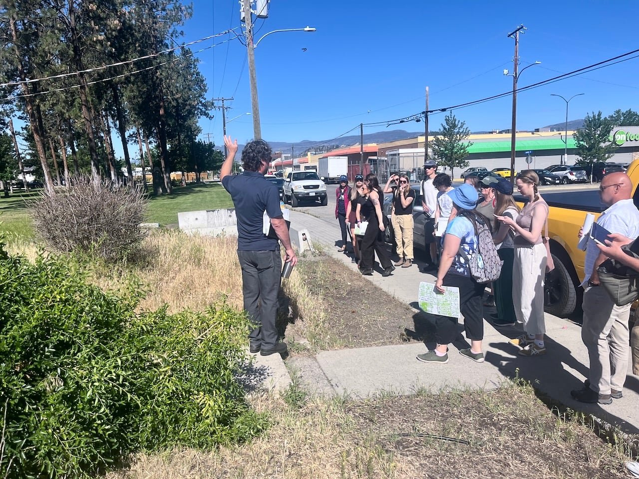 A man points to trees as he leads a group on a tour of a Kelowna neighborhood on a sunny day.