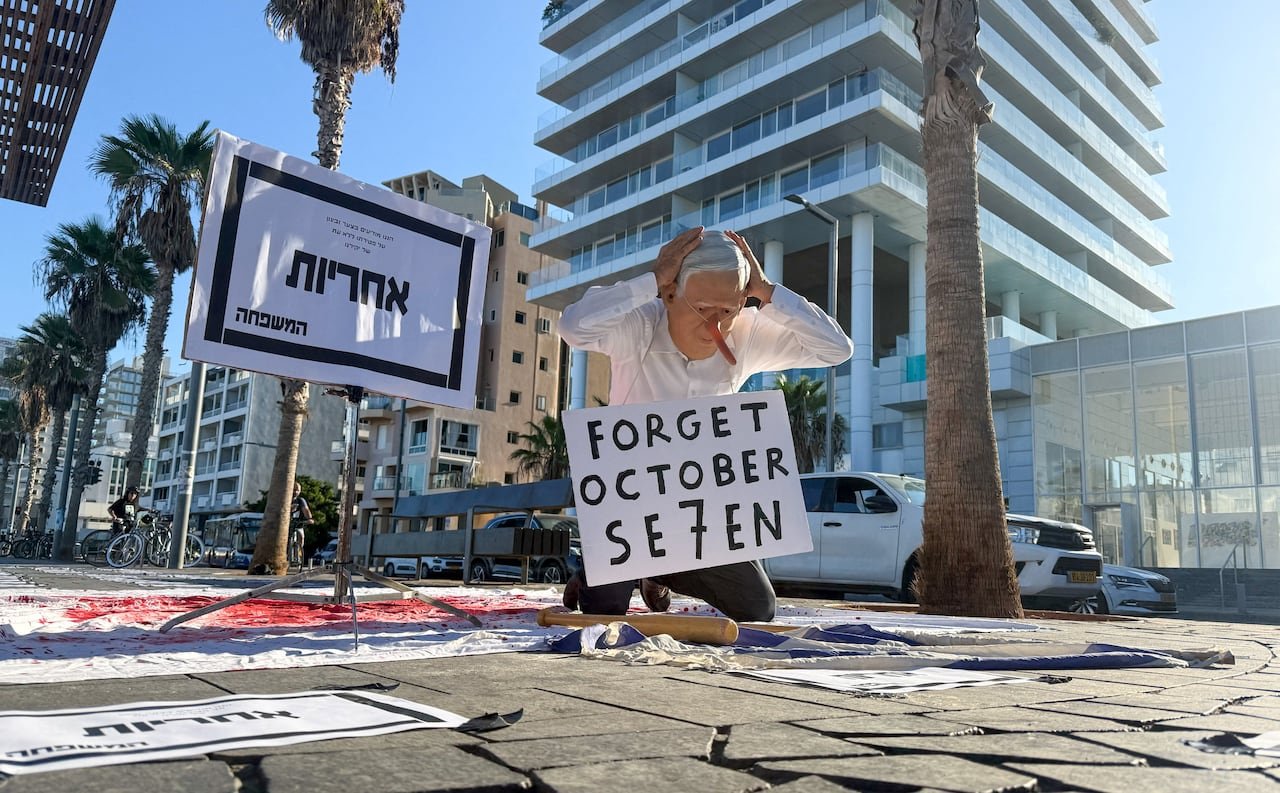 A protester dressed as Netanyahu kneels on a street with a sign around his neck that reads 'Forget October Seven' during the third anniversary of the attacks in Tel Aviv.