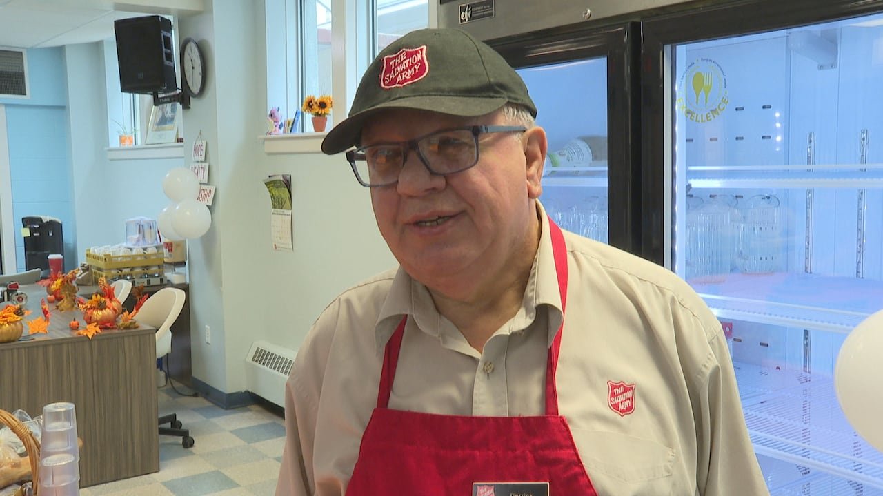 default-7876 a man wearing glasses, a Salvation Army baseball cap, and a red apron inside a facility next to a refrigerator.